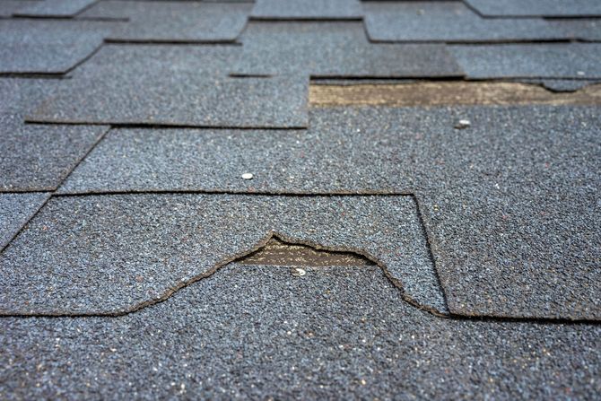 Two roofers, wearing hard hats, work on a black metal roof, one using a power drill.