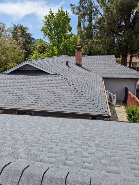Gray asphalt shingle roof on a house with a brick chimney, surrounded by trees on a sunny day.