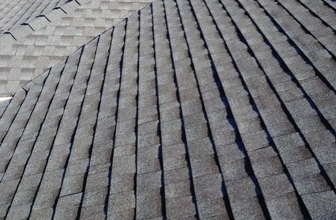 Two roofers, wearing hard hats, work on a black metal roof, one using a power drill.