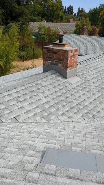 Brick chimney on a gray shingle roof, surrounded by trees and a sunny sky.