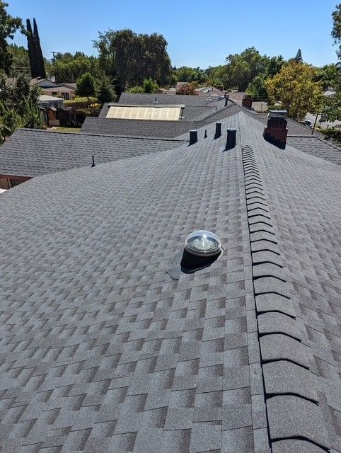 Gray asphalt shingle roof on a house with ventilation domes and vents, under a bright blue sky.