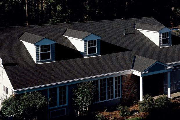 Two roofers, wearing hard hats, work on a black metal roof, one using a power drill.