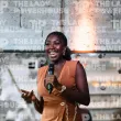 A smiling Black woman speaking into a microphone at a conference. She wears a tan dress in front of a brick background.
