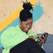 Woman with dreadlocks, glasses, and green shirt smiles while using her phone, set against a wood and paint-swatch backdrop.
