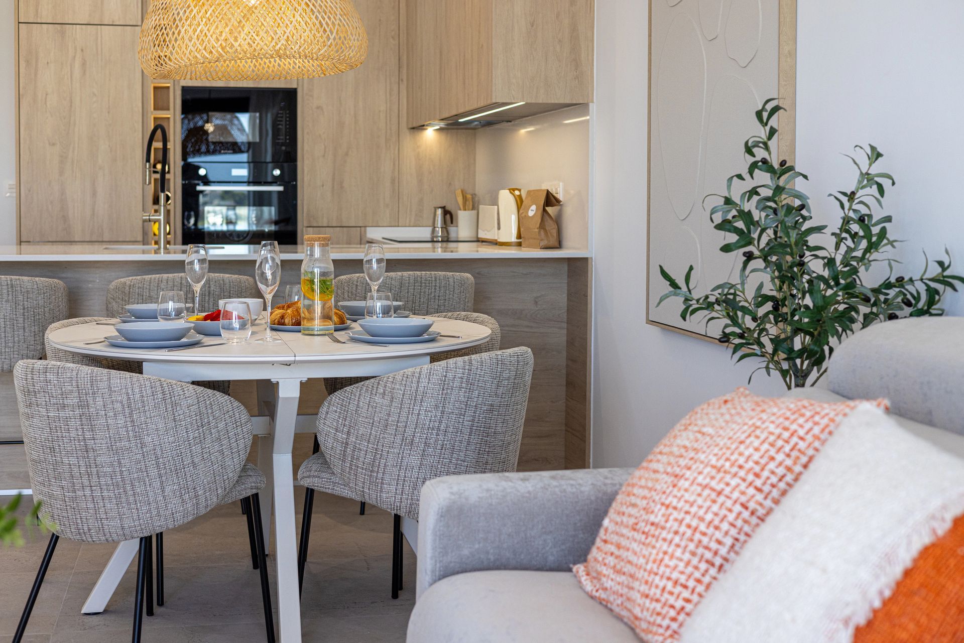 Zona pranzo con tavolo apparecchiato per i pasti, sedie intrecciate e cucina a vista con mobili in legno chiaro.