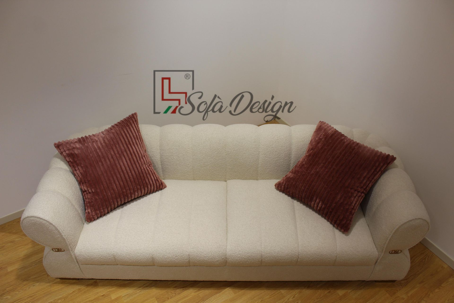 Divano beige con due cuscini bordeaux contro una parete bianca e un pavimento in legno. È visibile il logo Sofa Design.