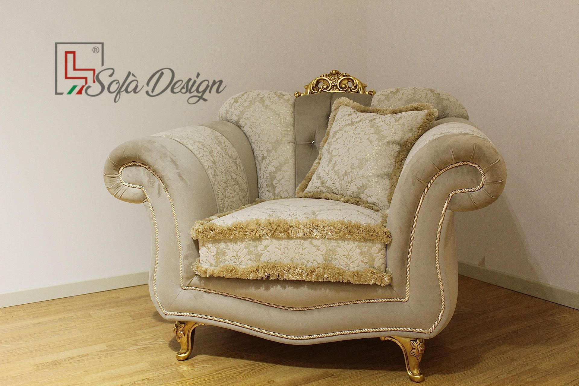 Poltrona decorata color crema con finiture dorate, frange e cuscino, appoggiata a una parete bianca in un angolo.