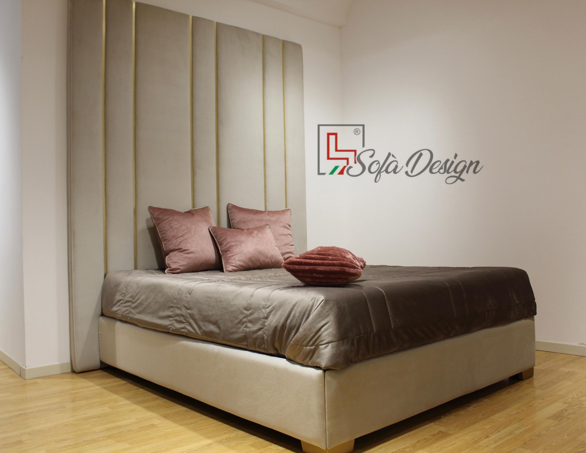 Letto con testiera alta imbottita beige e dettagli dorati, con cuscini marroni e rosa, su pavimento in legno.