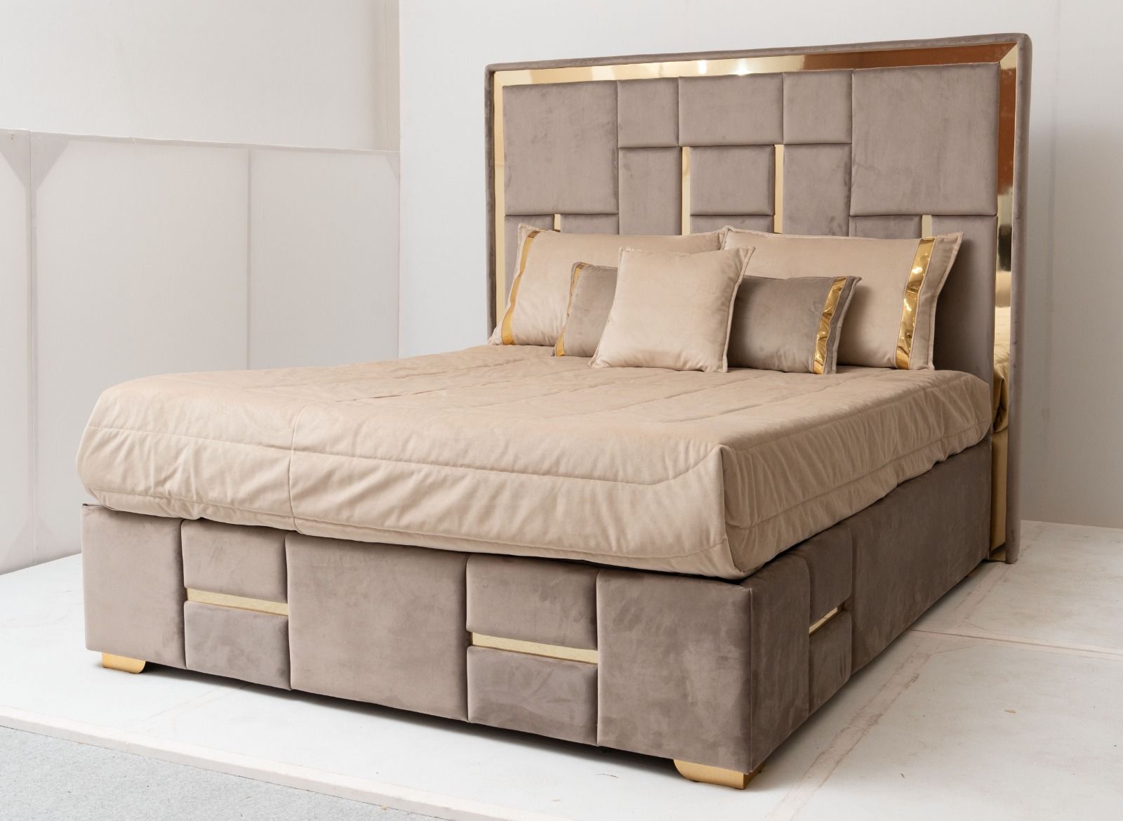 Letto imbottito con testiera, giroletto e base contenitore color tortora, con finiture dorate. Biancheria da letto beige con cuscini coordinati.