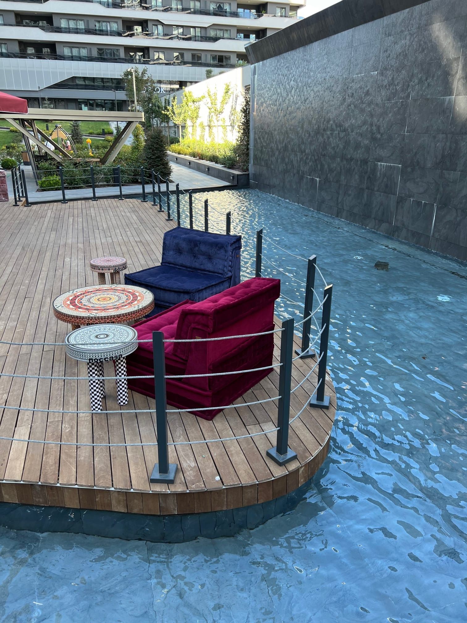 Area salotto all'aperto con sedie in velluto e tavoli rotondi su una terrazza in legno circondata dall'acqua.