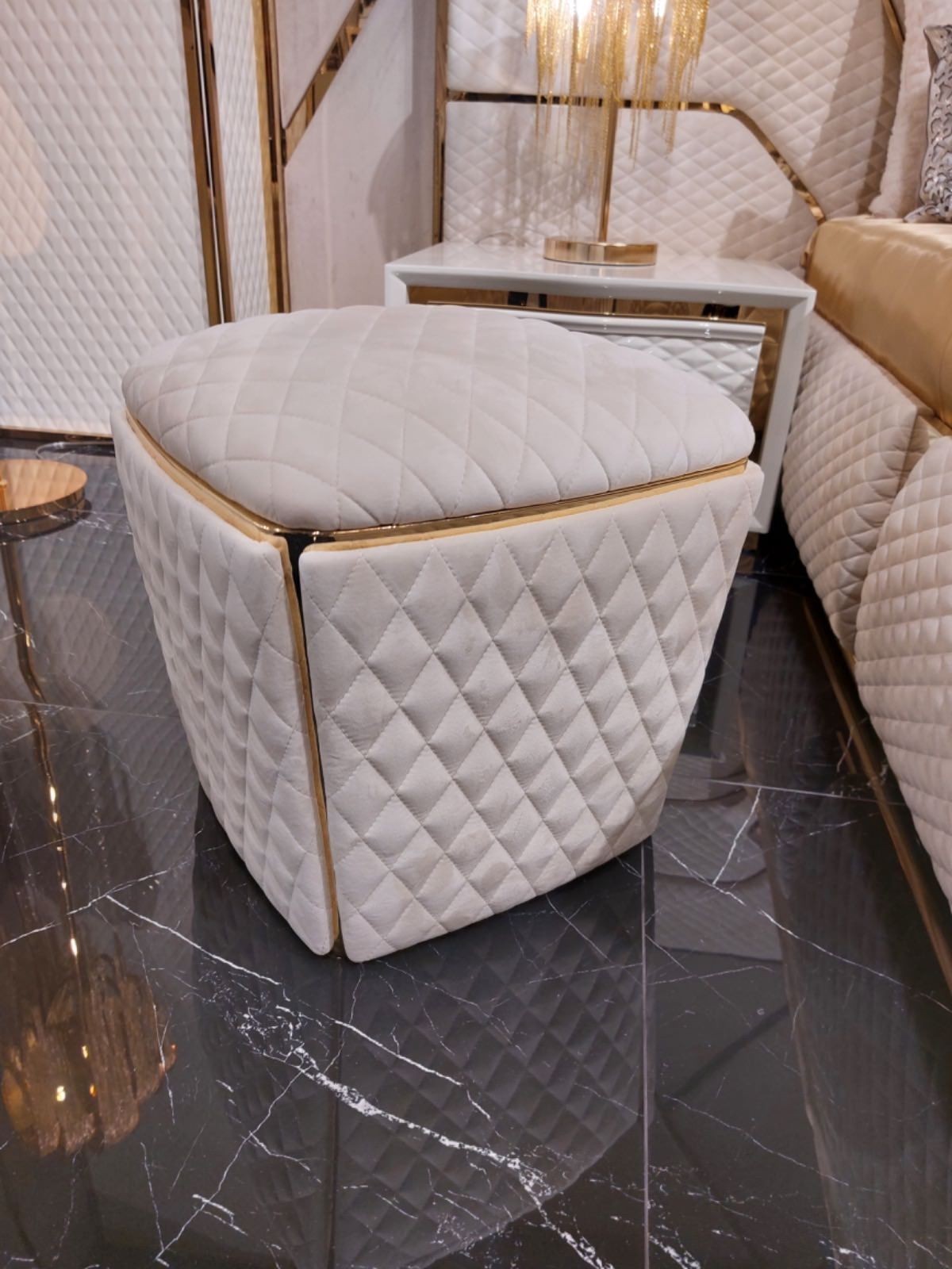 Pouf trapuntato color bianco sporco con finiture dorate, su un pavimento in marmo, accanto a un letto e a un comodino.