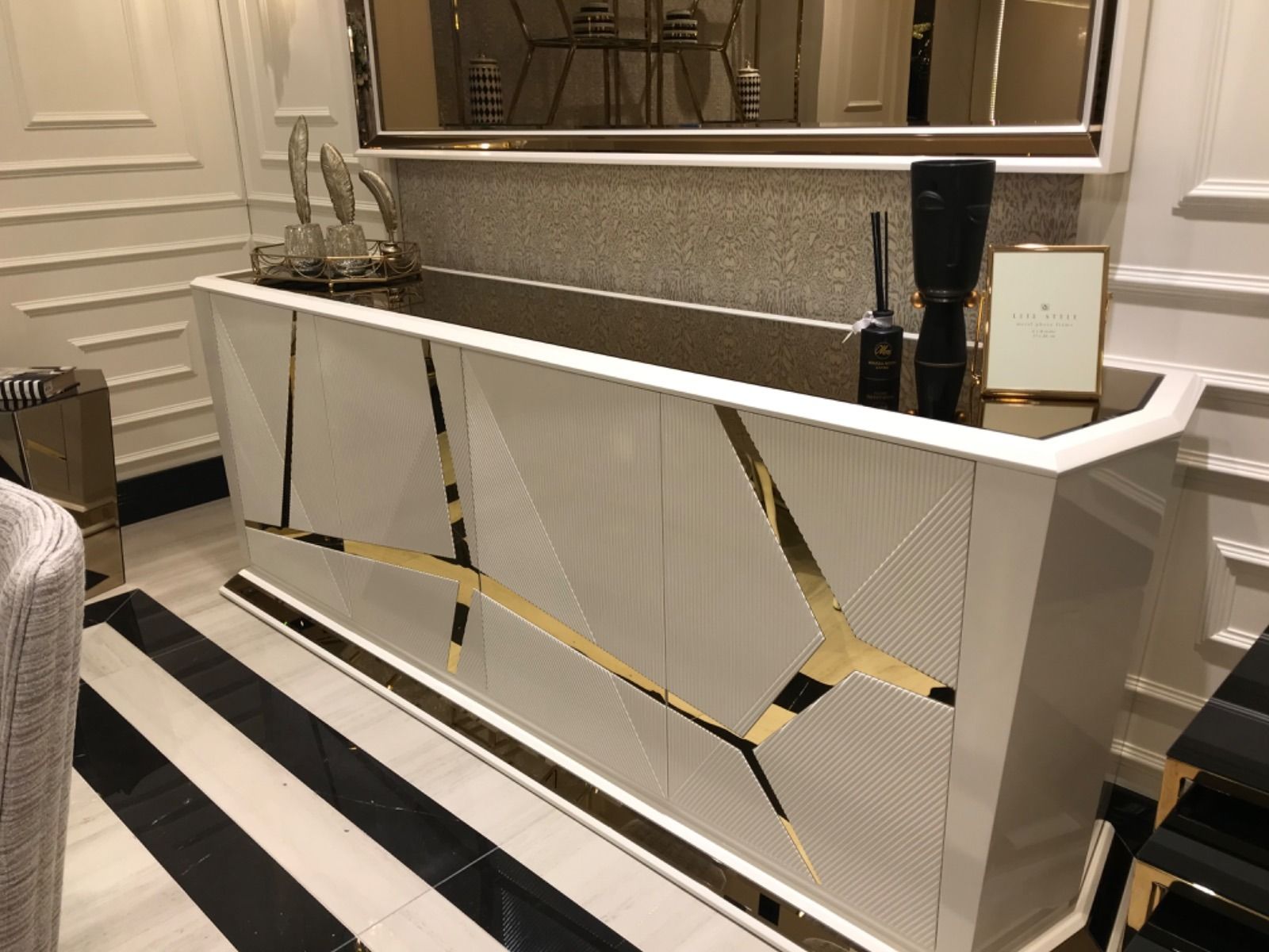Mobile bianco con dettagli dorati, inserito in una stanza con uno specchio decorativo e un tappeto a righe bianche e nere.