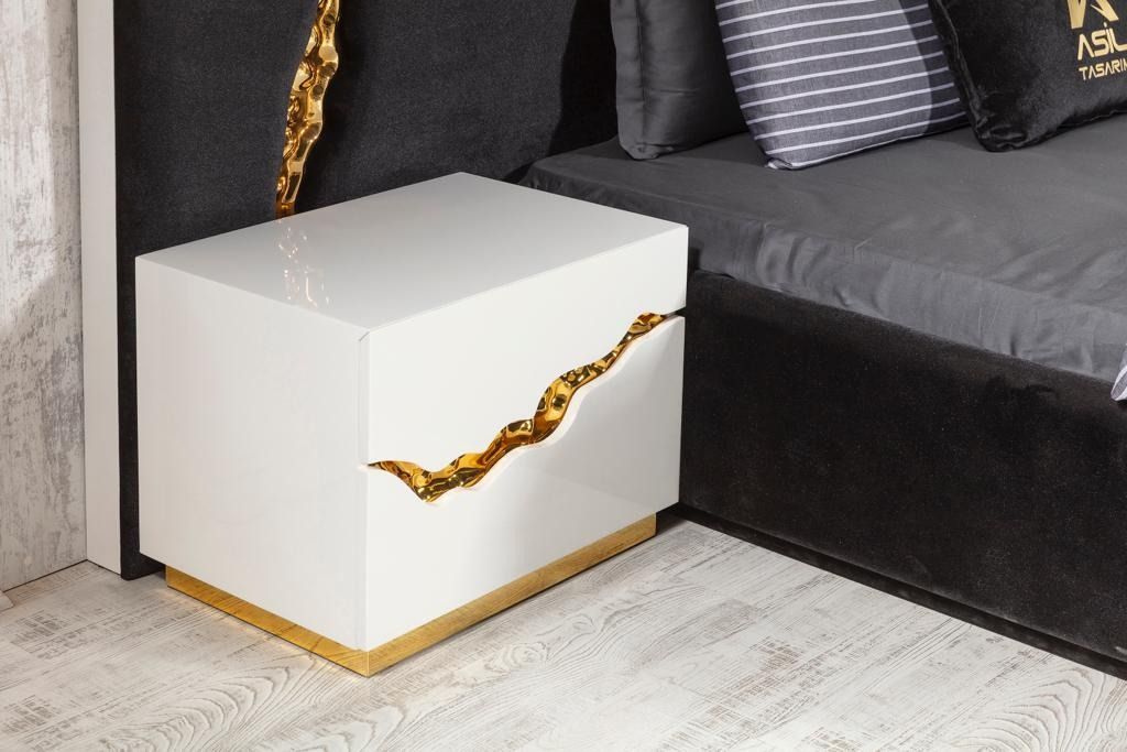 Comodino bianco con dettagli dorati accanto a un letto con cuscini scuri.