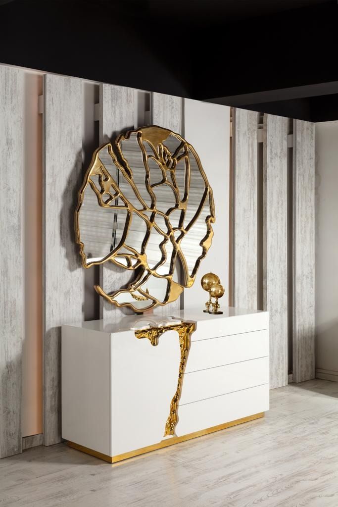 Comò moderno bianco con dettagli dorati, sotto uno specchio artistico con cornice dorata, contro una parete bianca testurizzata.