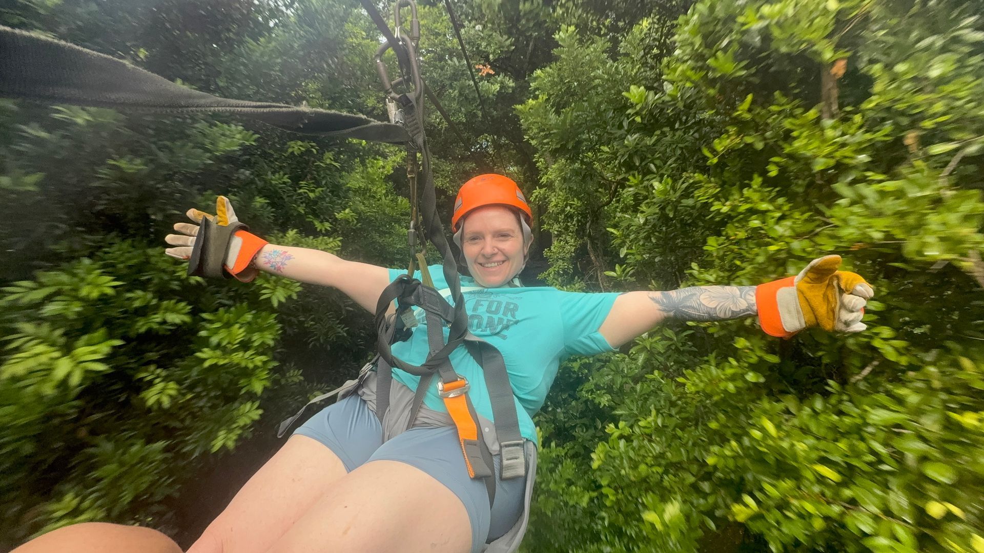 Person ziplining through a lush, green forest with arms outstretched, wearing a helmet and harness.