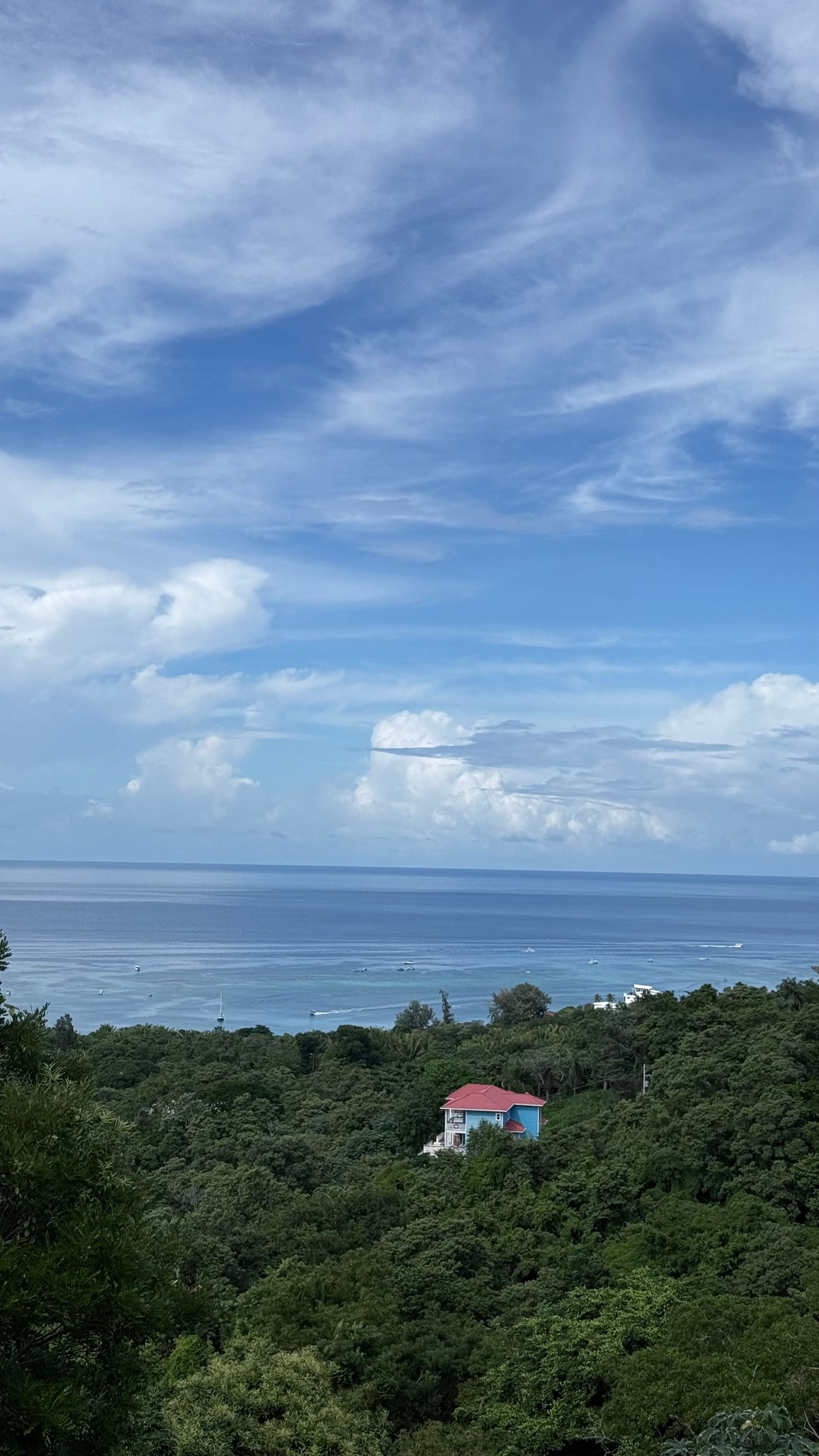 Blue sky with clouds over the ocean, with a house nestled in green trees.