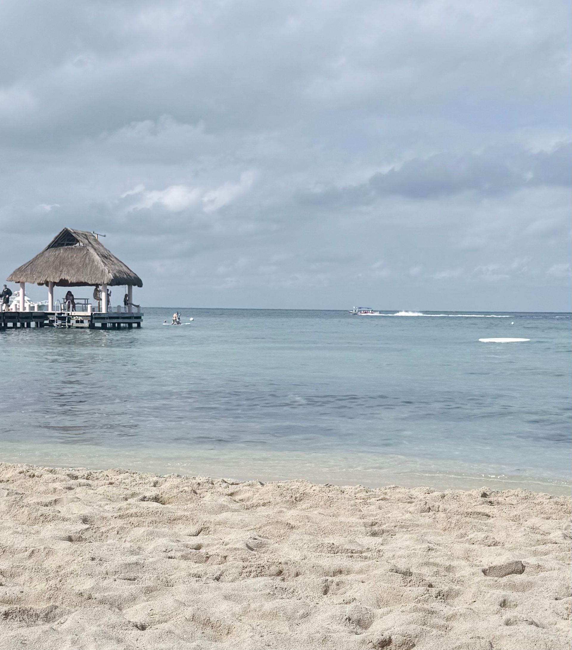 Beach scene with a thatched-roof gazebo on a pier. Calm, clear water; cloudy sky.