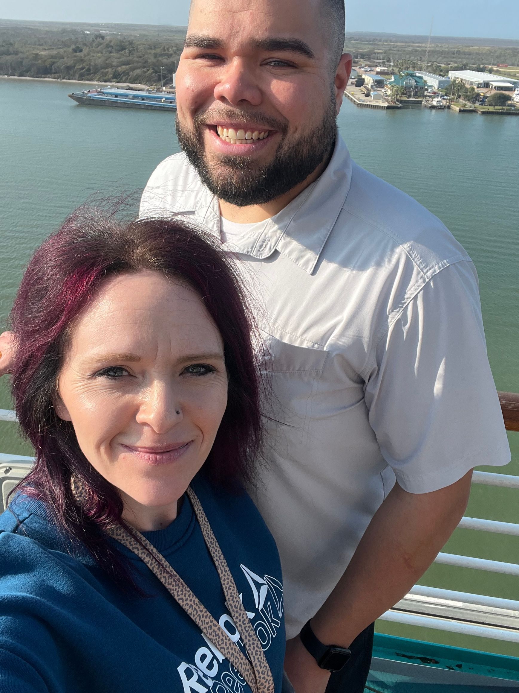 Couple smiling on a boat deck with a harbor view; the woman has purple-streaked hair.