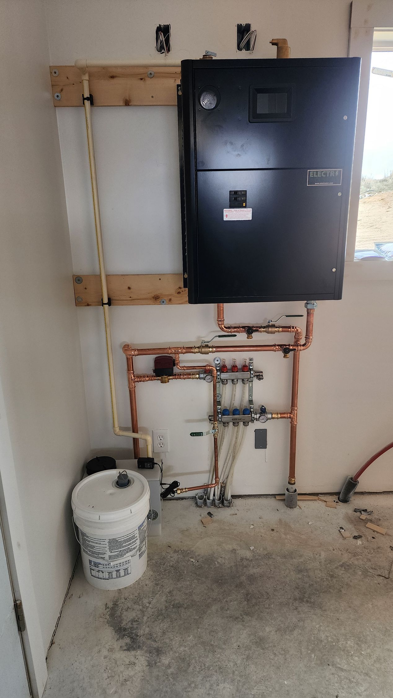 Heating system with black boiler, copper pipes, white tank, and a bucket in a corner.