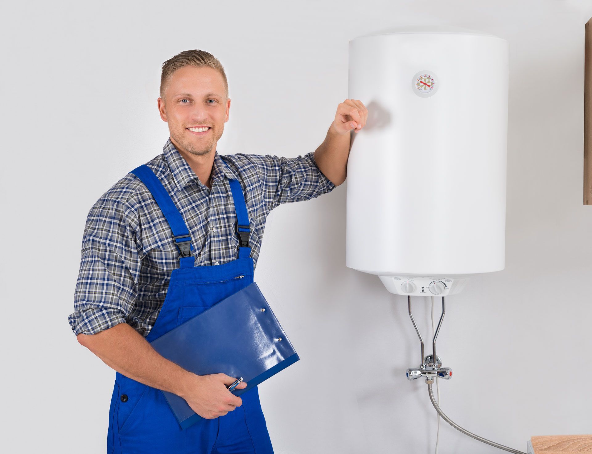 A man in blue overalls is holding a clipboard in front of a water heater.