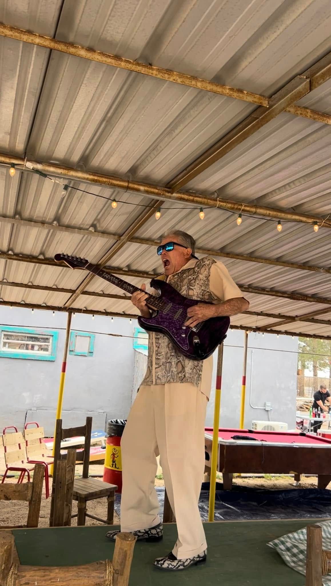Person playing a wind instrument under a metal-roofed pavilion, wearing a patterned shirt and white pants.