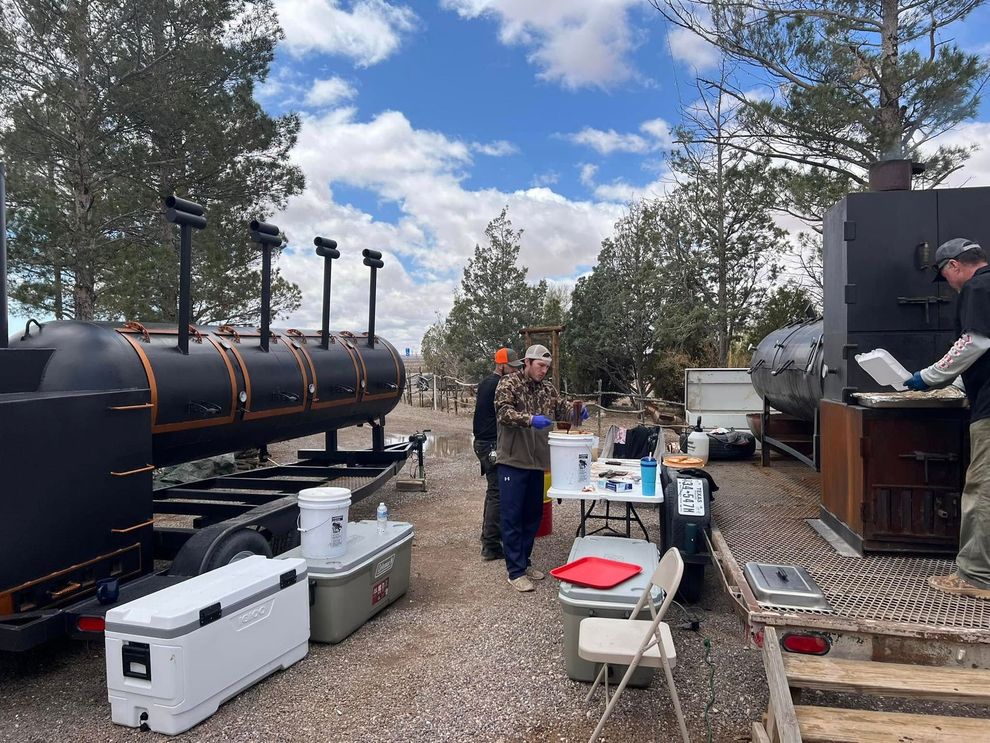 Outdoor barbecue setup with a black smoker, folding tables, and a person cooking beside it.