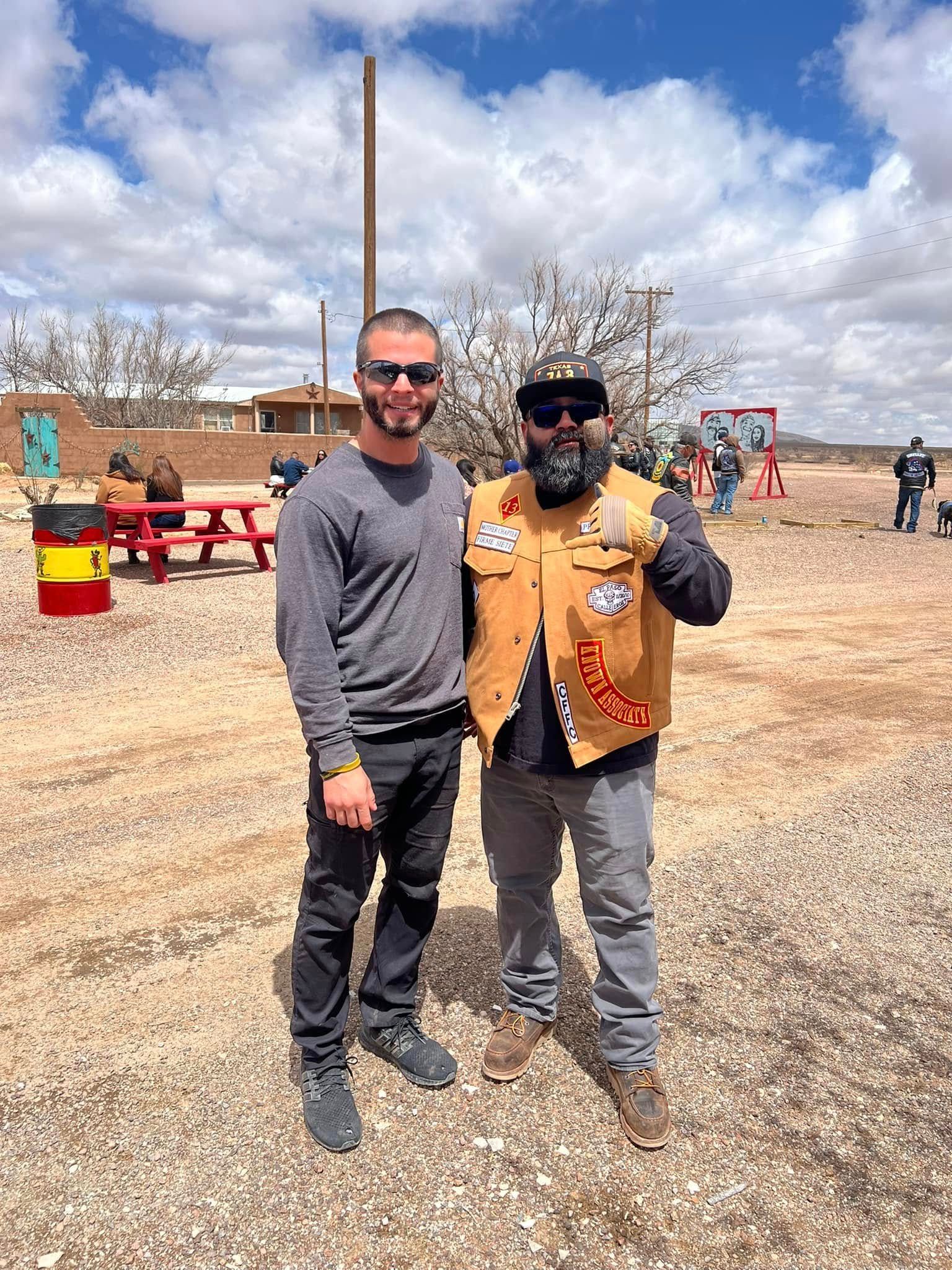 Two men posing outdoors at a desert event, one in a gray shirt and one in a tan vest giving a thumbs-up.