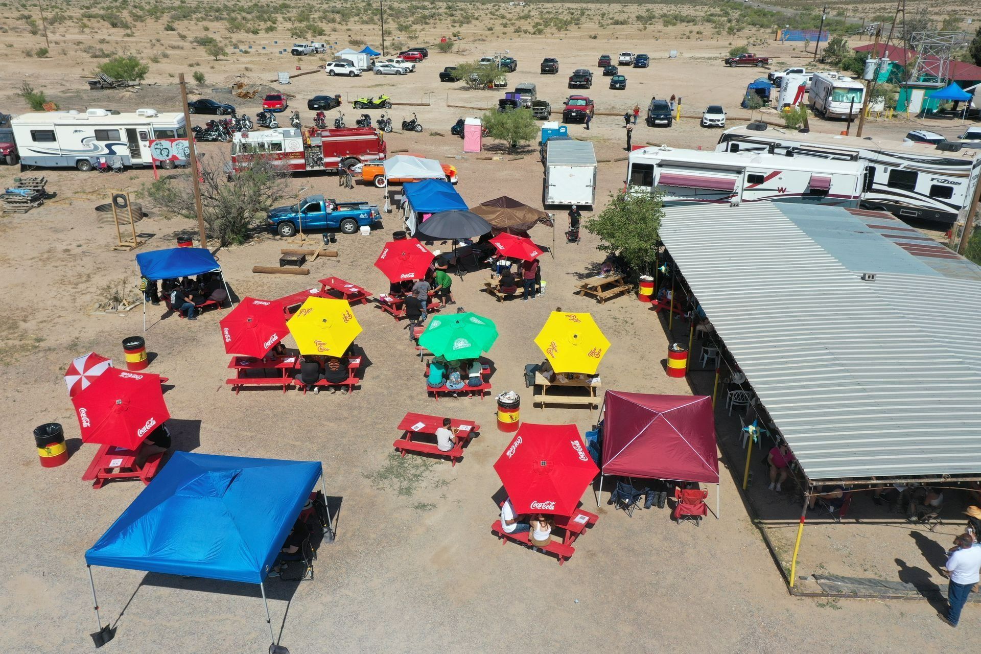 Aerial view of a desert camp with colorful tents, RVs, and tables arranged around a large canopy.
