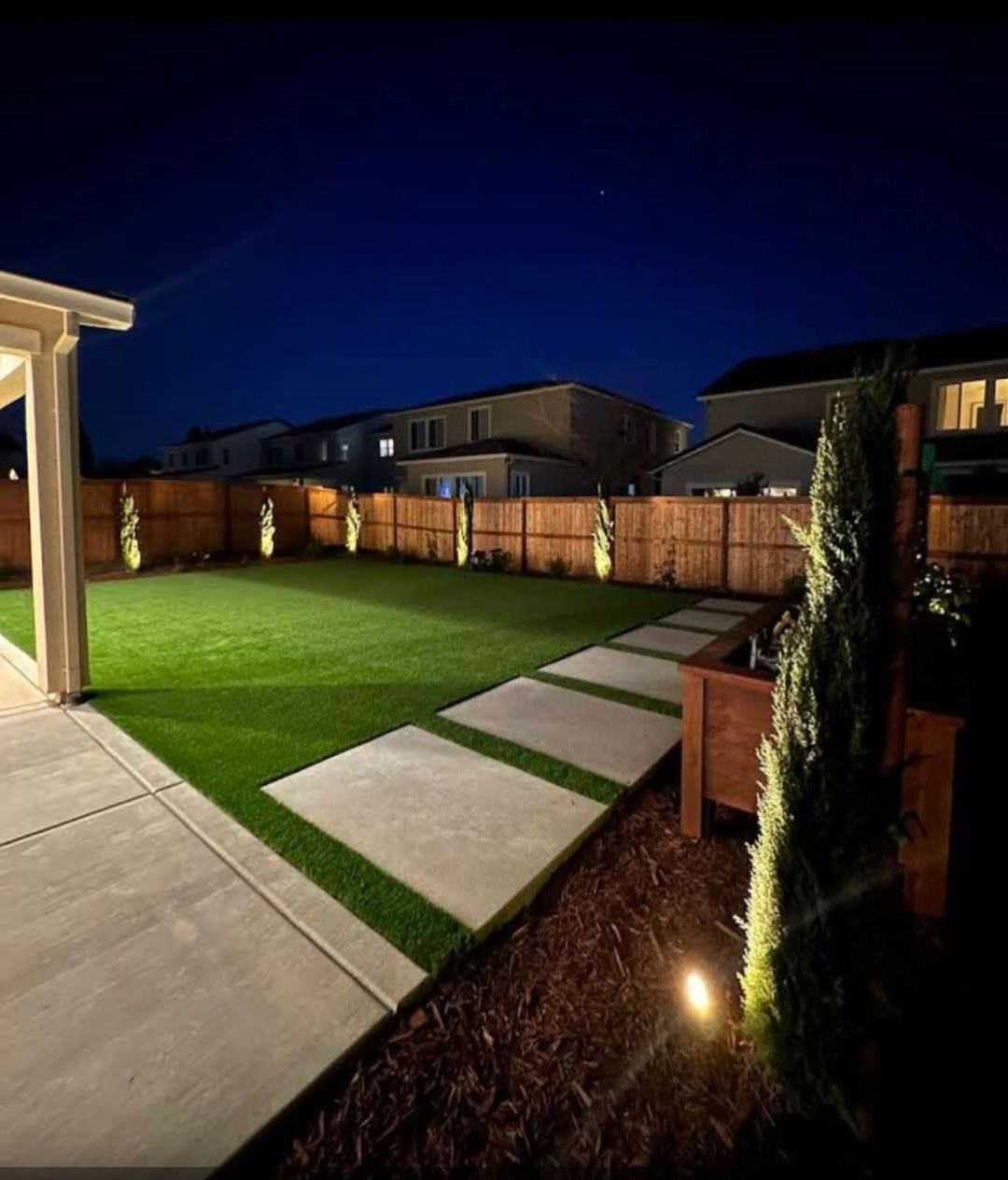 Well-lit backyard at night with a green lawn, concrete path, and decorative lights.