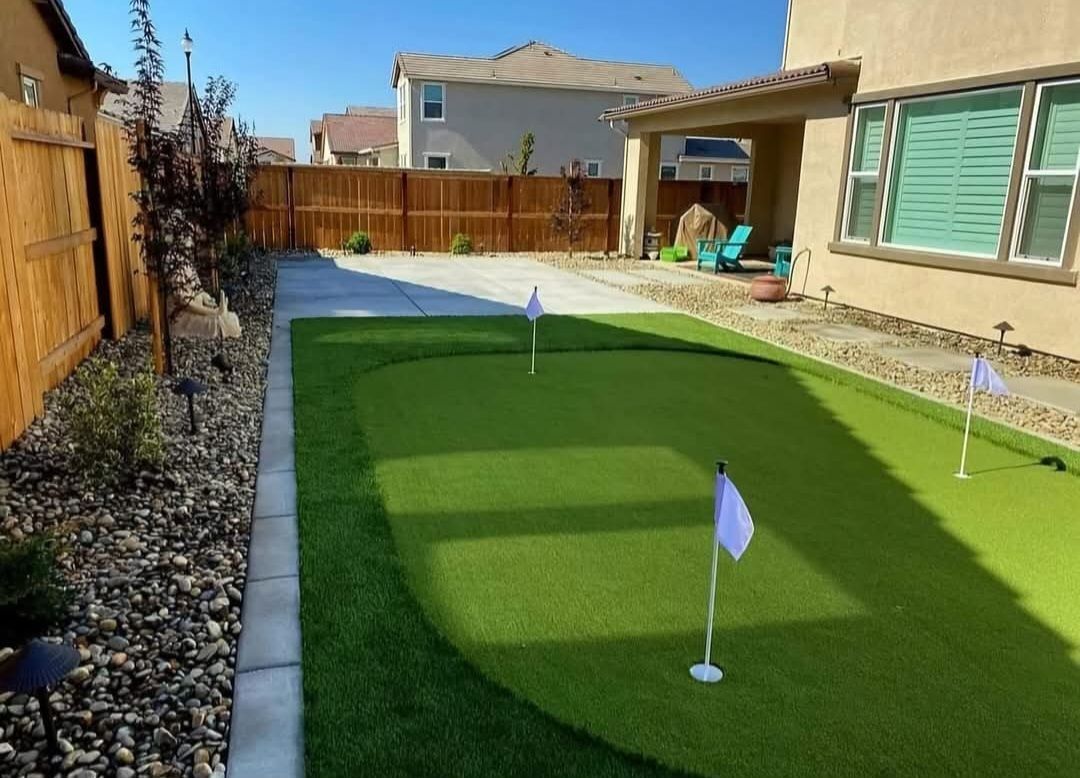 Backyard with artificial putting green, flags, and stone border. House and fence in background.