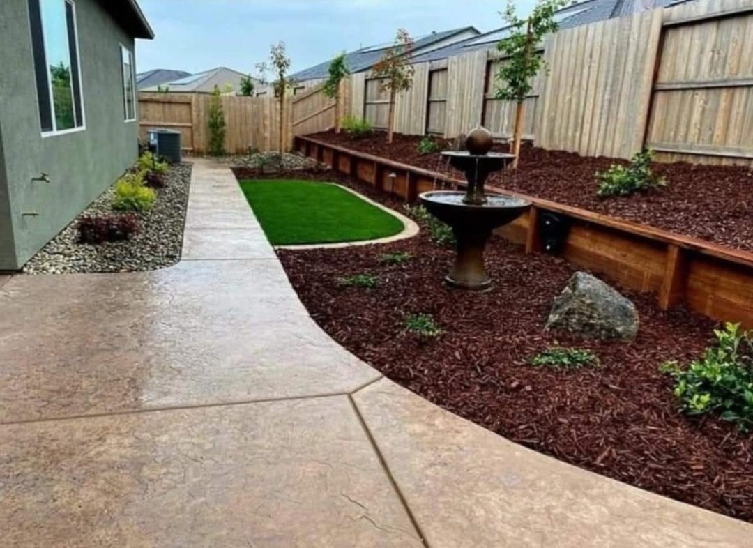 Backyard with concrete pathway, fountain, artificial turf, and mulch beds.