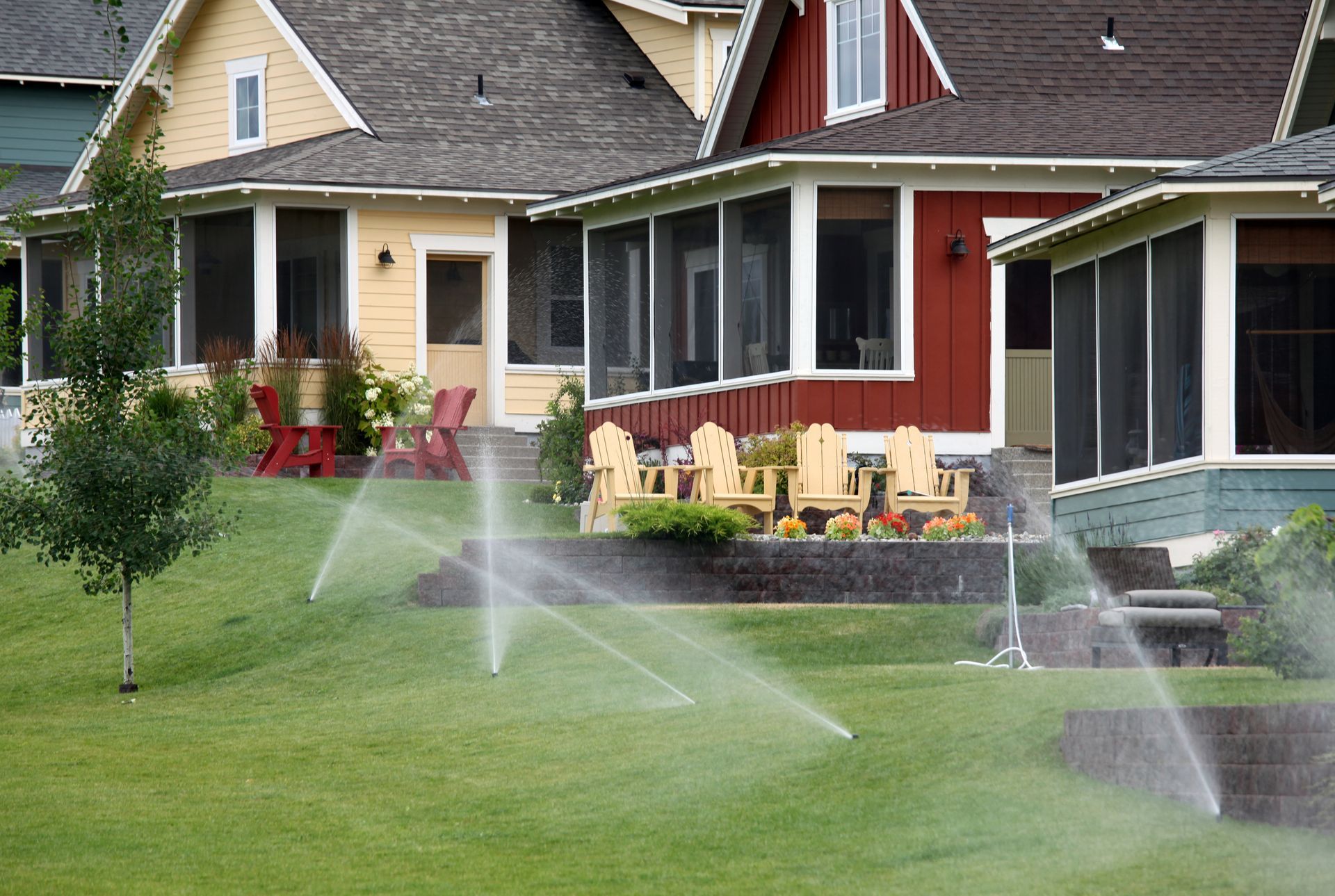 Lawn sprinklers spraying water on a green lawn in front of colorful houses with screened porches.