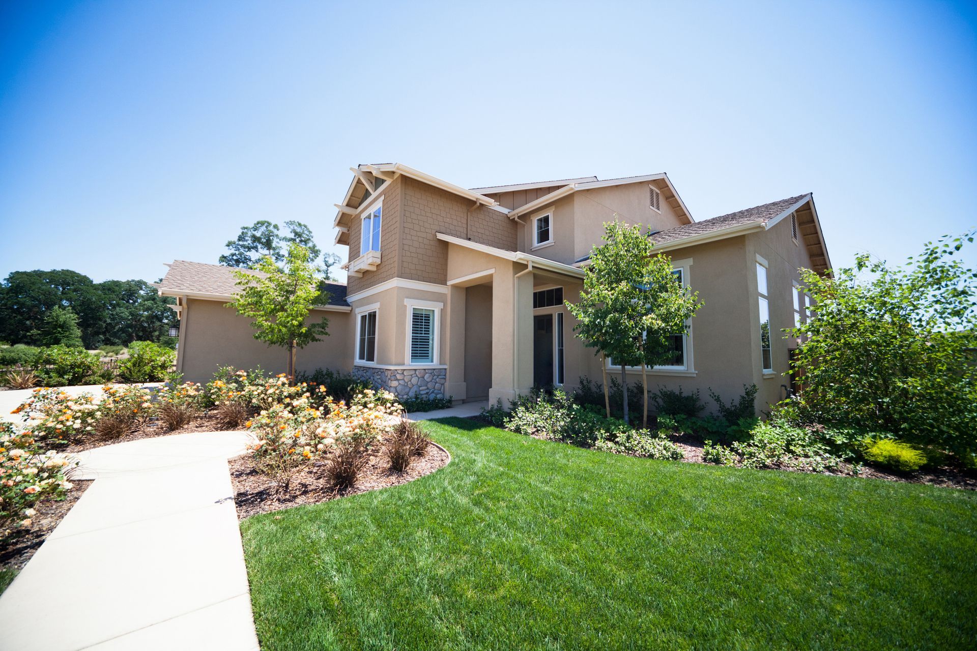 House with tan stucco exterior, green lawn, blue sky, and flowering plants in front.
