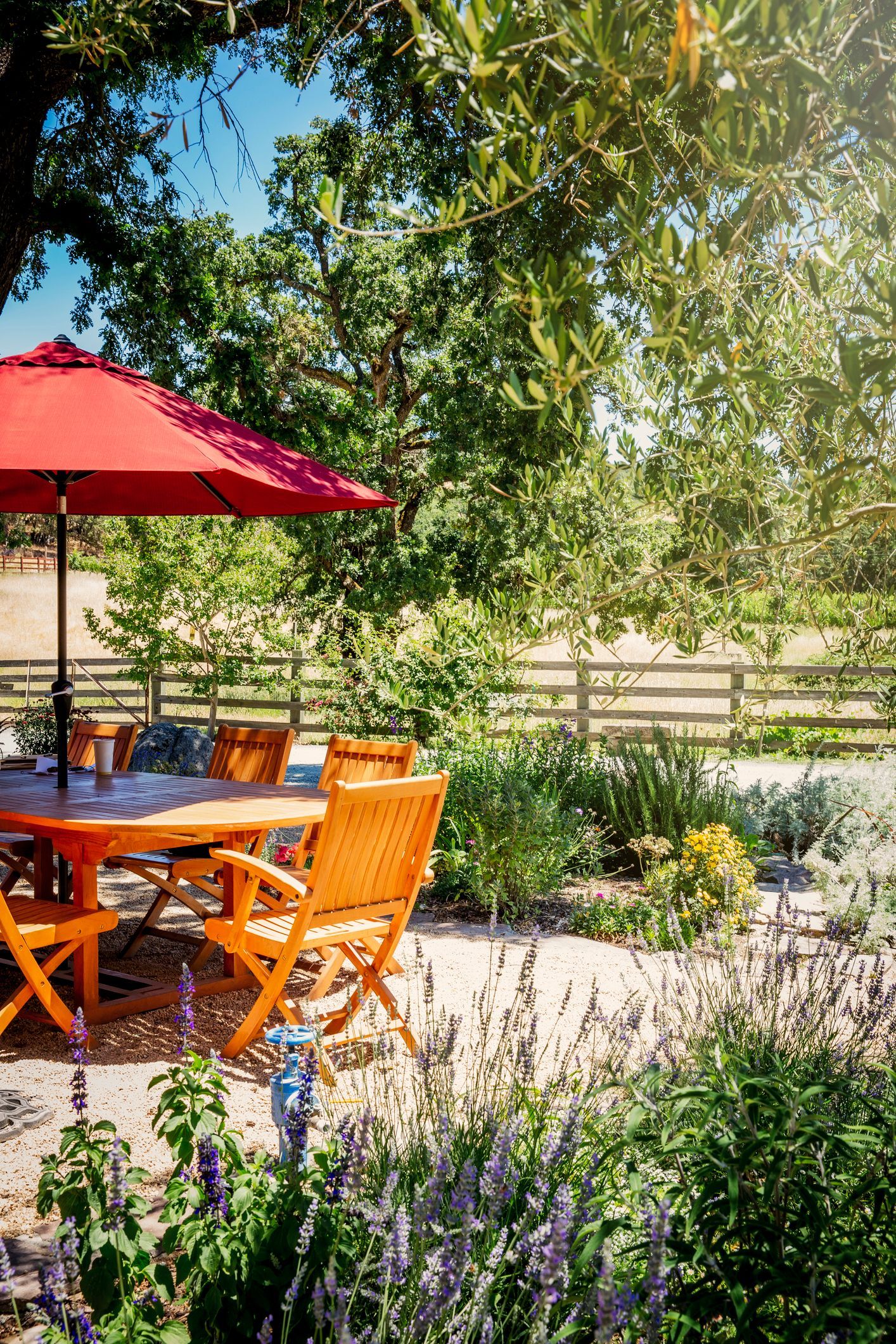 Outdoor patio with table and red umbrella, overlooking garden and fence.