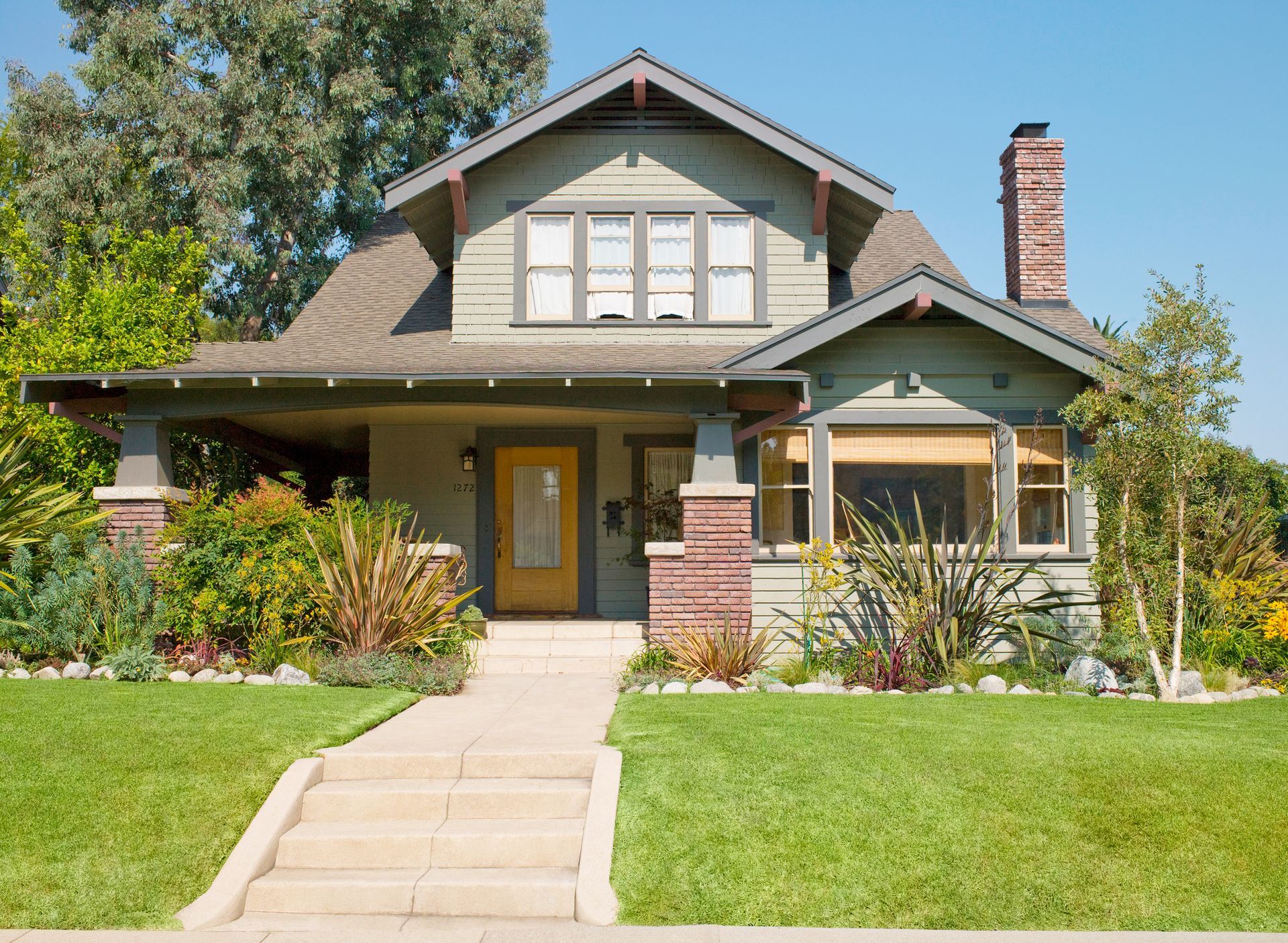 Green Craftsman home with porch, brick accents, yellow door, and sunny lawn.