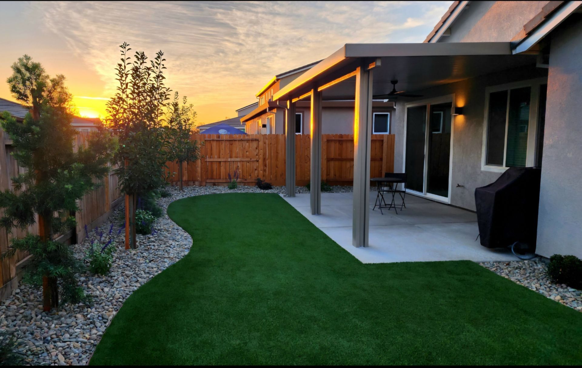Backyard landscaping with turf installed, concrete patio, concrete walkways, a patio cover installed and rock installed in the planters with the plants and trees.
