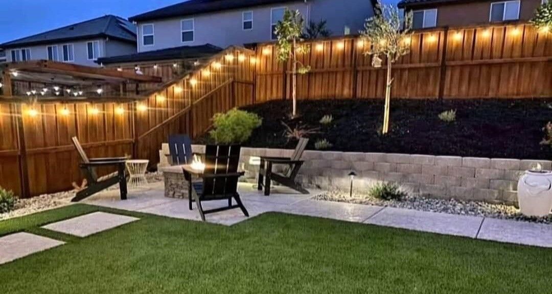 Backyard with string lights, fire pit, seating, and a raised garden bed against a fence.