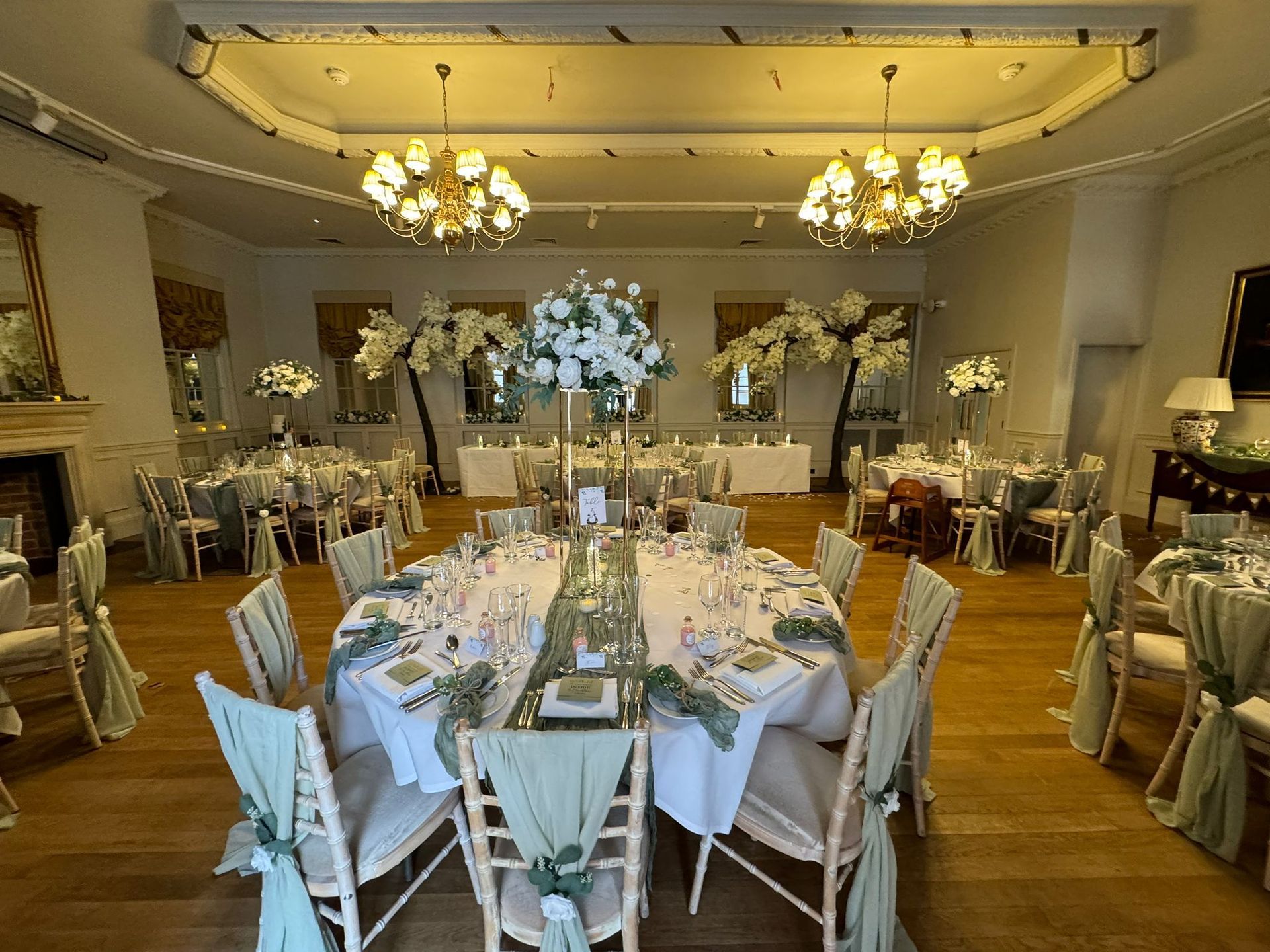 Wedding reception: round tables, centerpieces, chairs with green fabric, gold chandeliers in a ballroom.