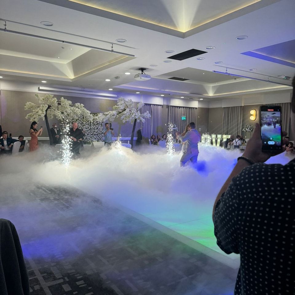 Wedding reception with fog on the dance floor; people are dancing. Decorative trees and lighting.