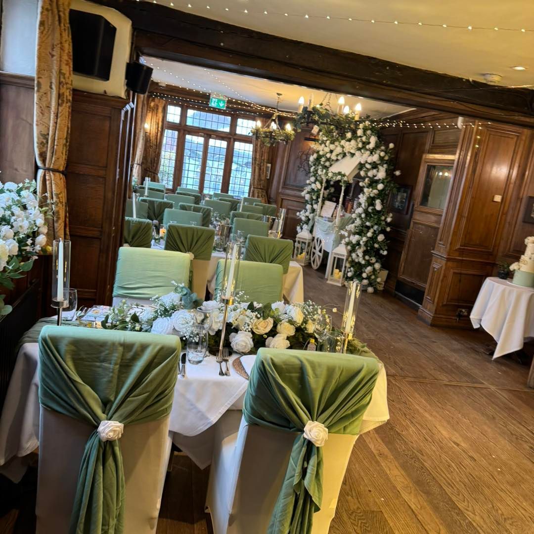 Wedding reception with tables, chairs decorated with sage green sashes, white floral arrangements, and a white archway.