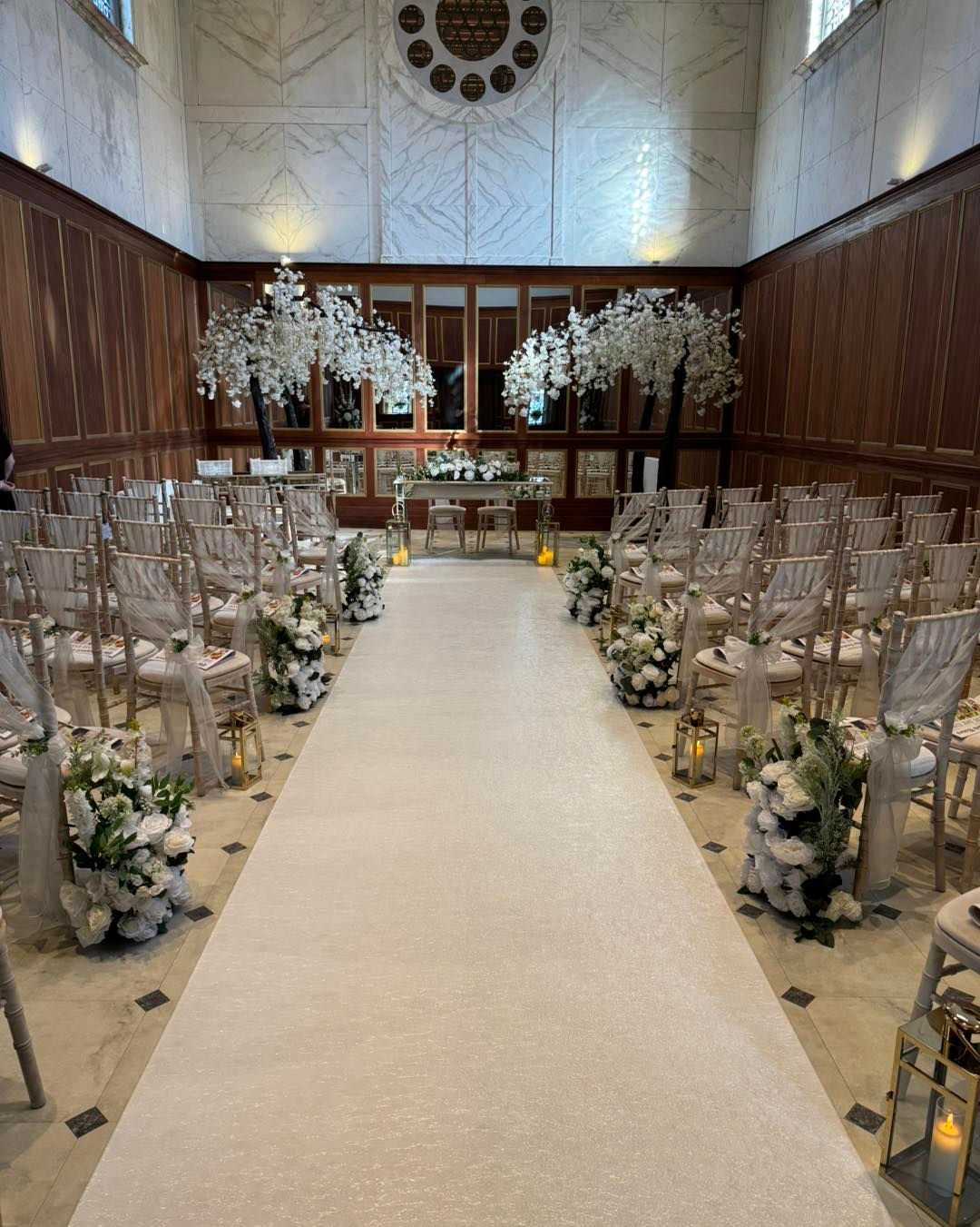 Wedding ceremony setup: aisle with flower arrangements, gold chairs, and floral backdrop in a wood-paneled room.