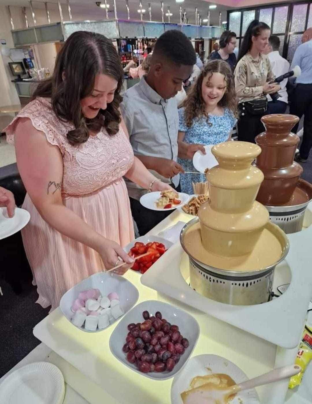 People at a buffet with chocolate fountains. Woman smiling, lad and girl behind. Bowls of snacks are on display.