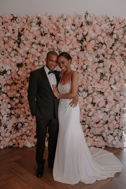 Newlyweds pose in front of a pink floral wall. The man wears a tuxedo, and the woman wears a wedding dress.