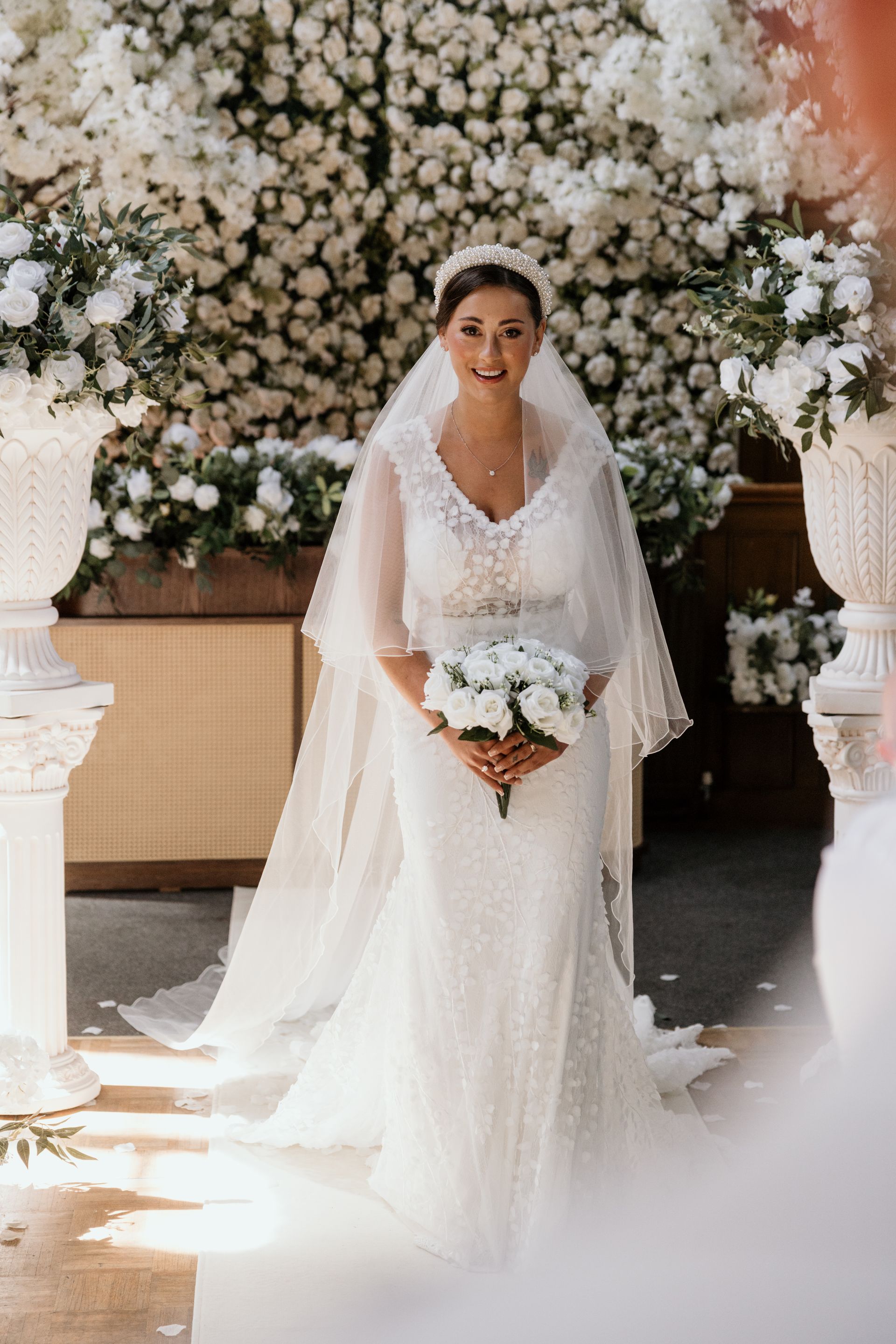 Bride in white gown holds a bouquet, standing in front of a flower wall and marble pillars.