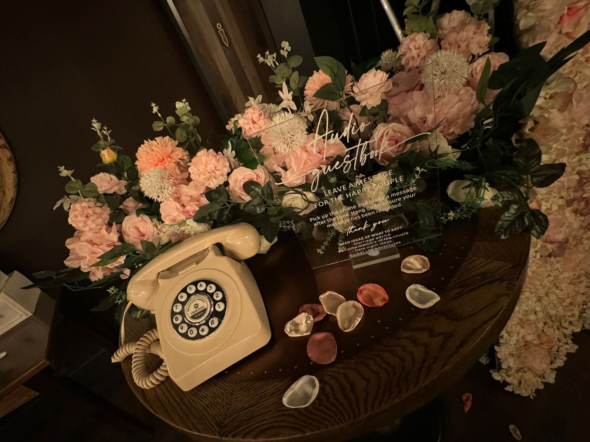 Cream rotary phone with pink flowers on a dark wooden table, scattered petals.
