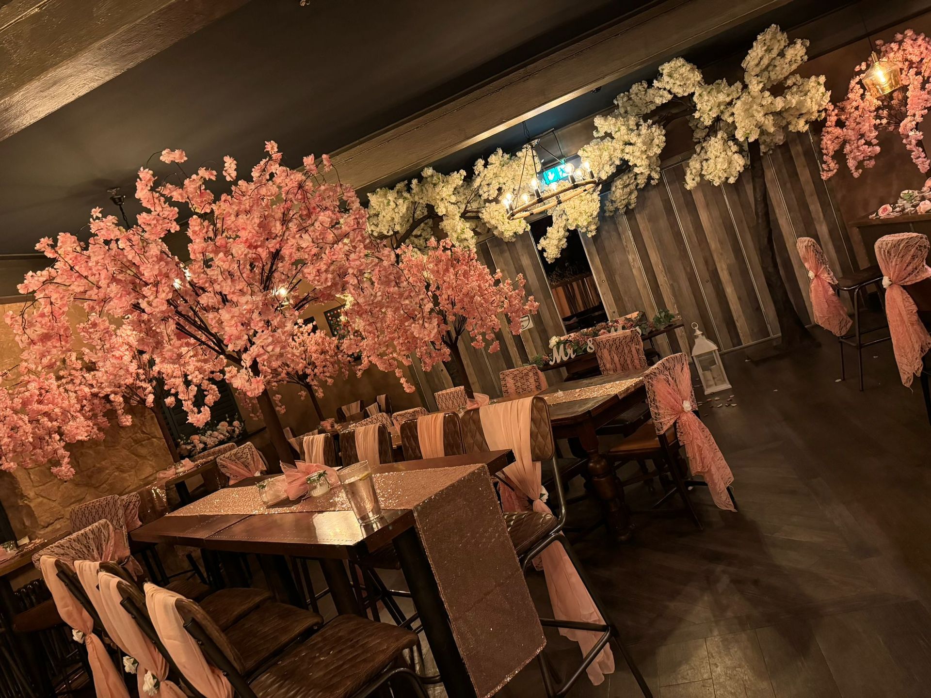 A dimly lit event space decorated with pink and white cherry blossom trees. Tables are set for guests.