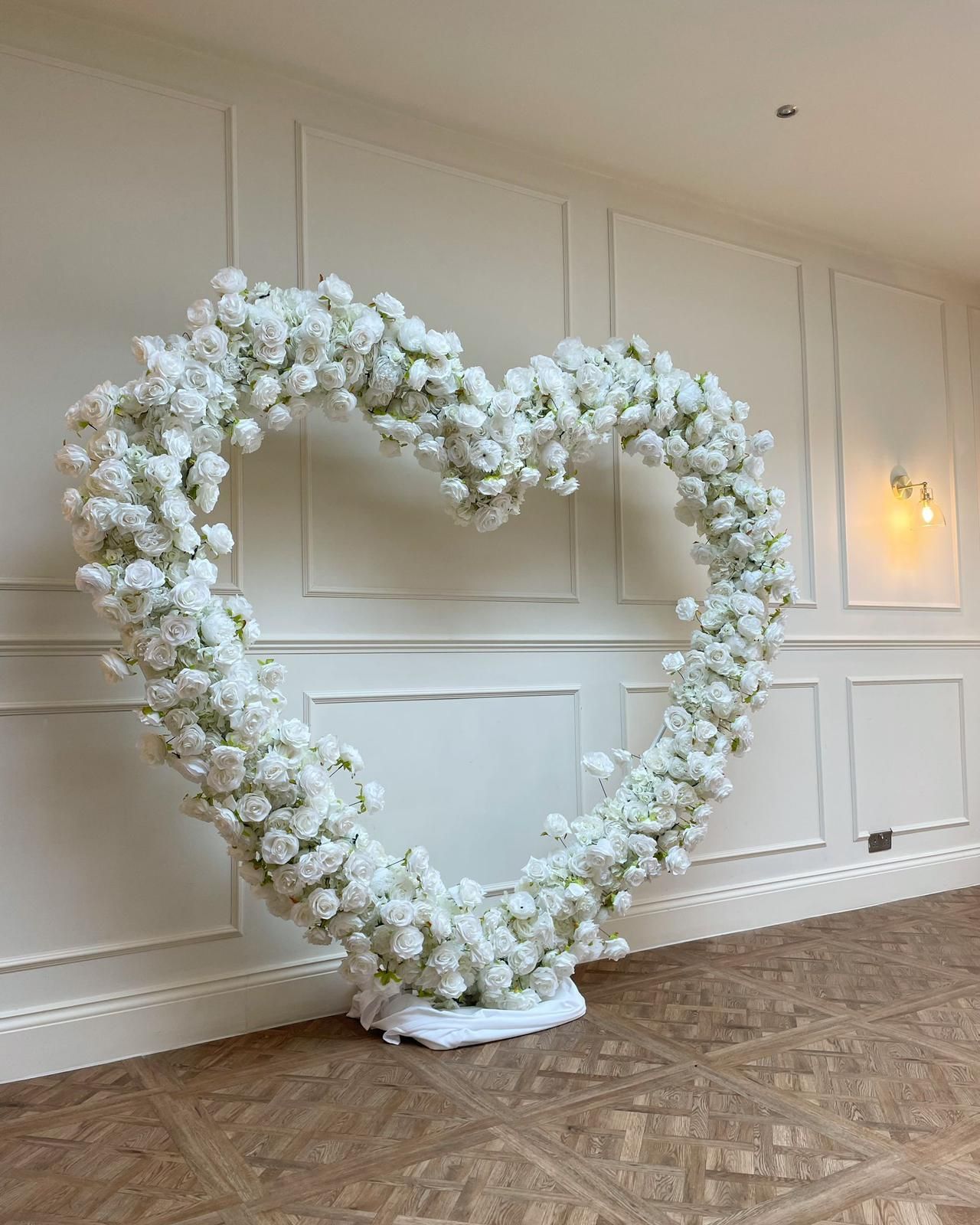 White floral heart archway on a white base, against a pale wall and wood floor.