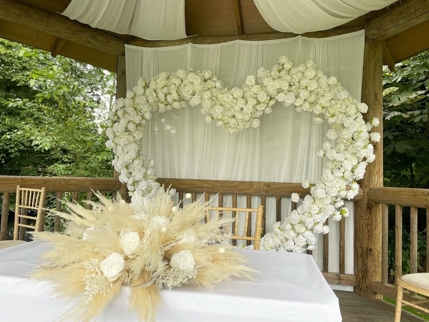 Wedding ceremony setup: white floral heart arch, decorated gazebo, white tablecloth with pampas grass centerpiece.
