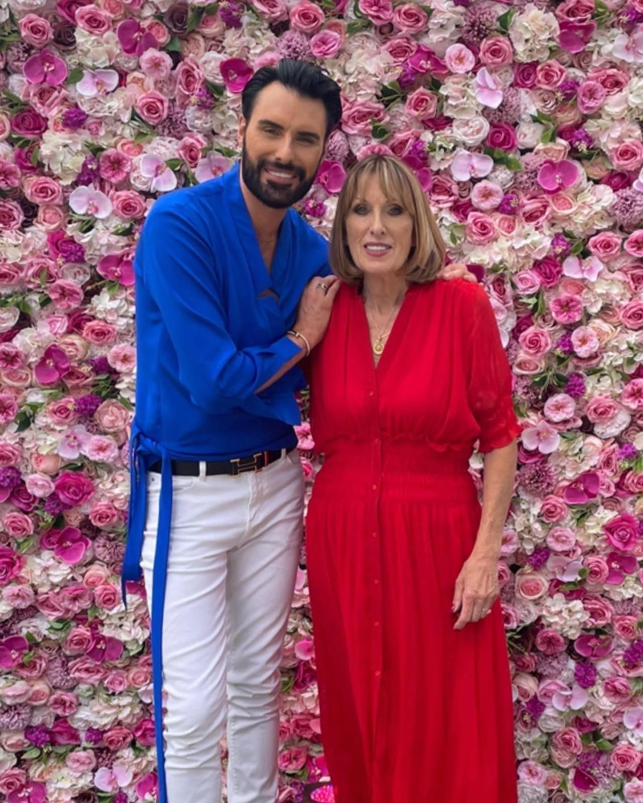 Rylan Clark in blue, and Lorraine Kelly in red, pose in front of a flower wall, smiling.