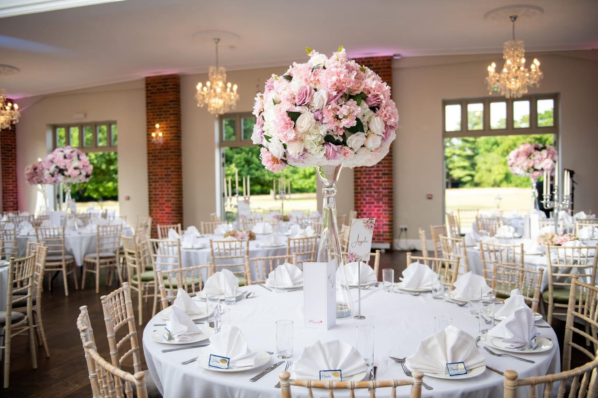 Elegant wedding reception with round tables, floral centerpieces, and chandeliers in a bright room.